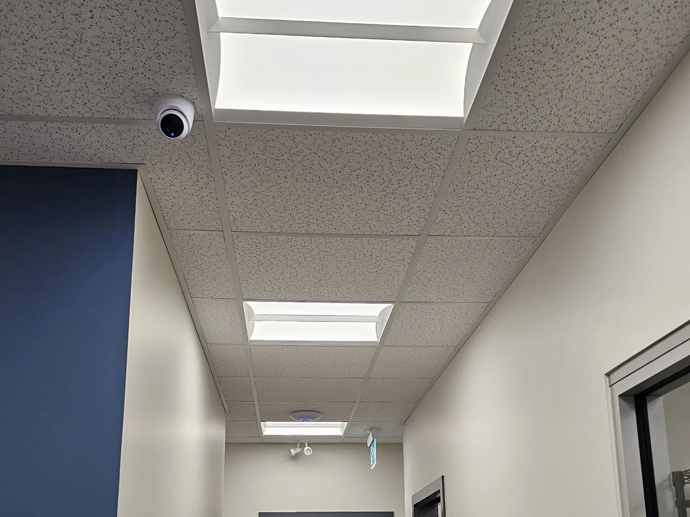 An office corridor installed by KeenAI with a ceiling-mounted dome camera, recessed LED troffers and a ceiling speaker