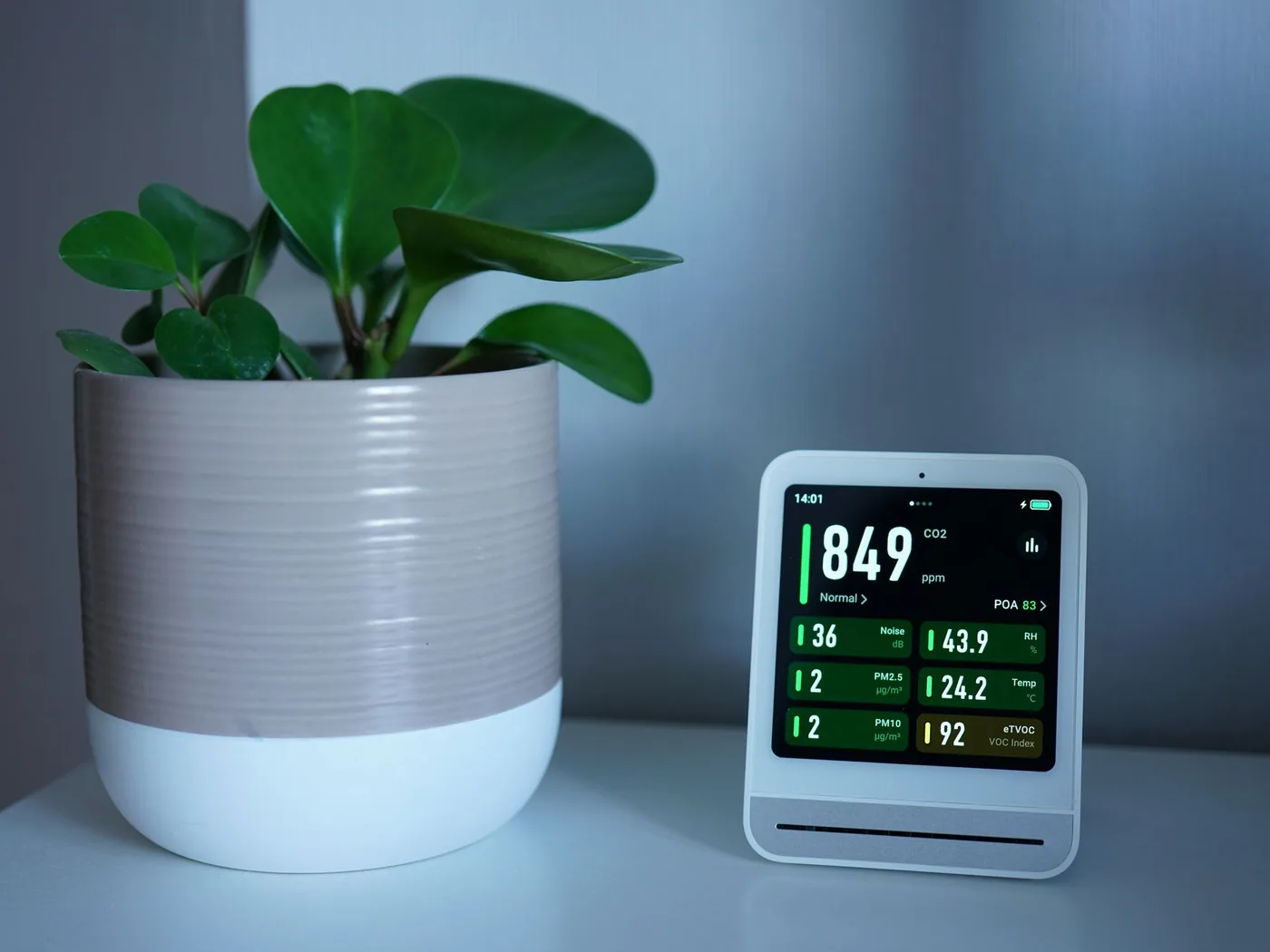 A desktop indoor air quality monitor beside a houseplant, displaying carbon dioxide in parts per million alongside fine particulate, humidity, temperature and volatile organic compound readings