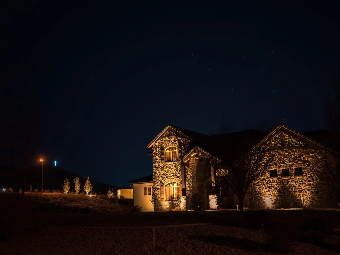 A stone-faced house after dark, lit by ground-mounted uplights on the facade with low path lights along the driveway and uplit trees